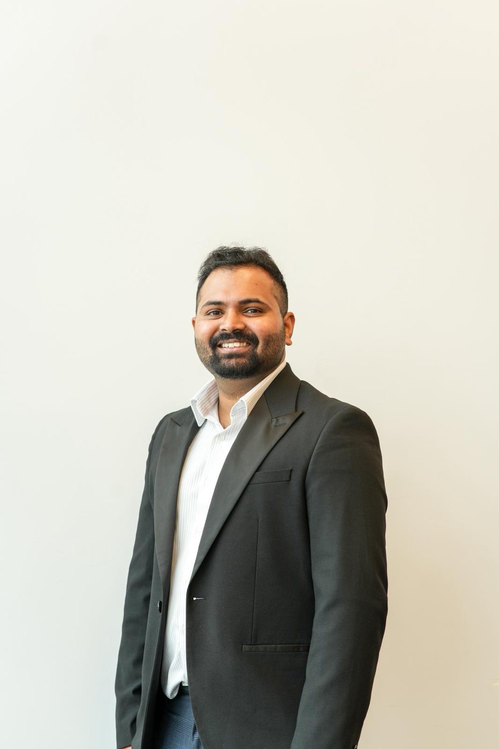 Thomas Kunjumon, smiling, in a dark jacket and white shirt against a plain light wall
